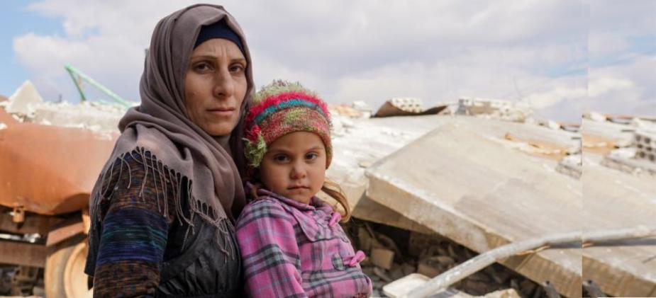 A woman holding a young child amid collapsed building rubble in Syria.