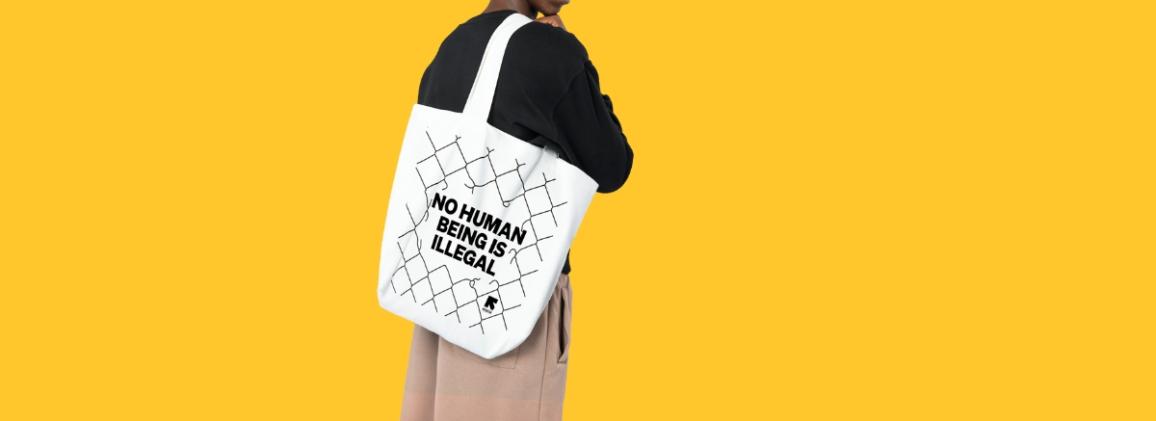 Beige tote bag with long handles featuring a black chain-link fence illustration with the text 'No human being is illegal' on the front in the center.