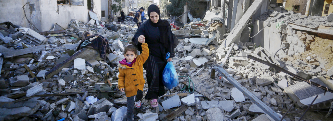 A woman leads a young child by the hand through the rubble of destroyed buildings.