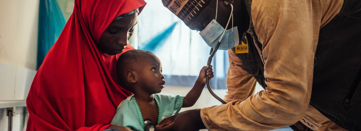 An IRC health worker examines a young child with a stethoscope while the mother holds the child in West Africa.