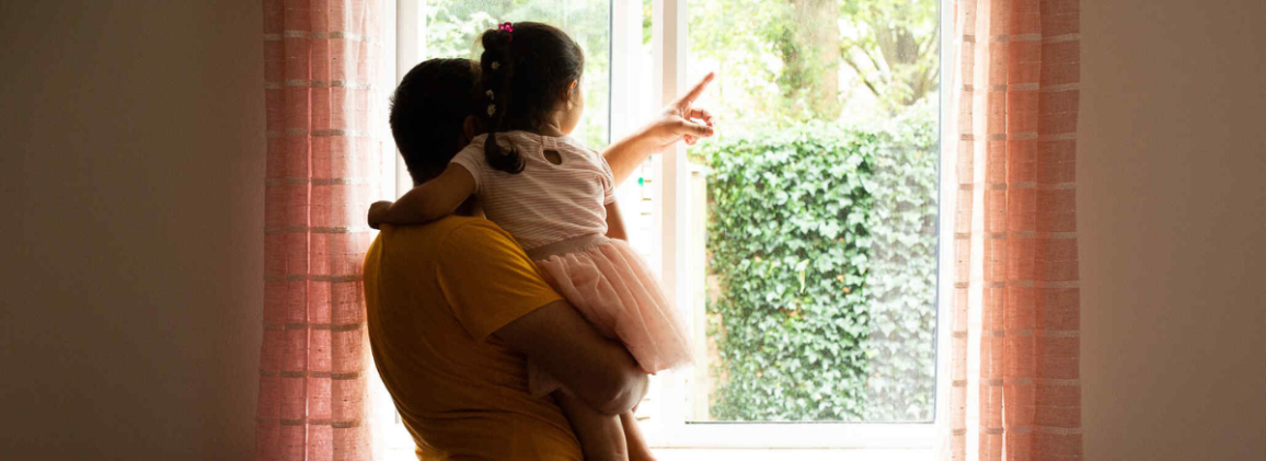 A parent holding a young child as they look out a window together at a garden.